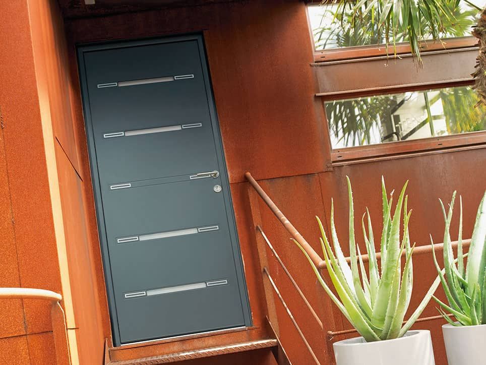 Porte extérieure gris foncé avec des inserts horizontaux en forme de fenêtre et une poignée argentée, adossée à un mur et un escalier de couleur rouille, avec des plantes en pot.