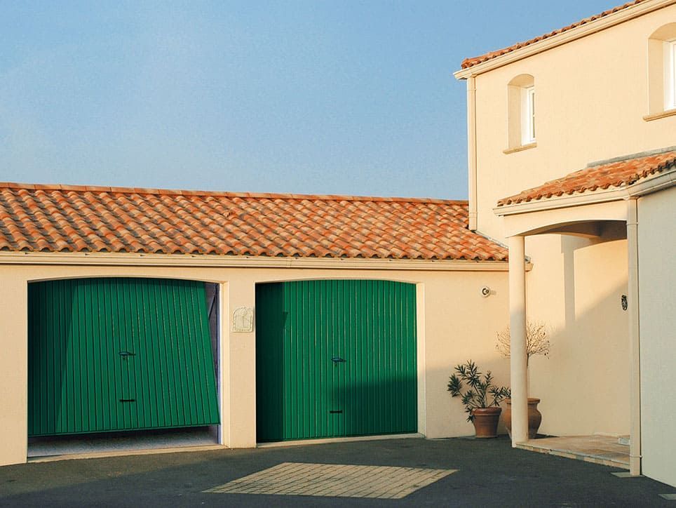 Deux portes de garage vertes sur un bâtiment de couleur claire, toit en terre cuite, ciel clair.