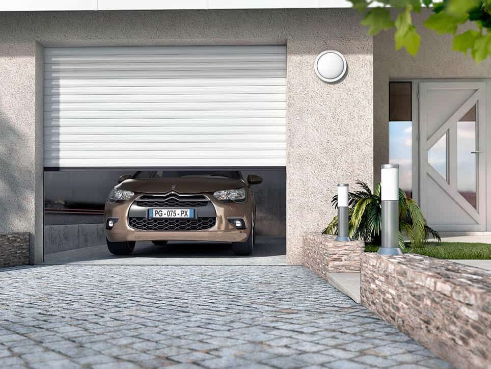 Voiture beige dans un garage avec porte enroulable blanche, à côté d'une porte d'entrée et d'une passerelle.