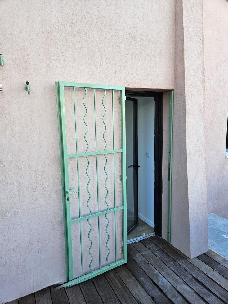 Porte de sécurité métallique verte ouverte devant une porte vitrée. Extérieur du bâtiment avec mur en stuc rose et terrasse en bois.