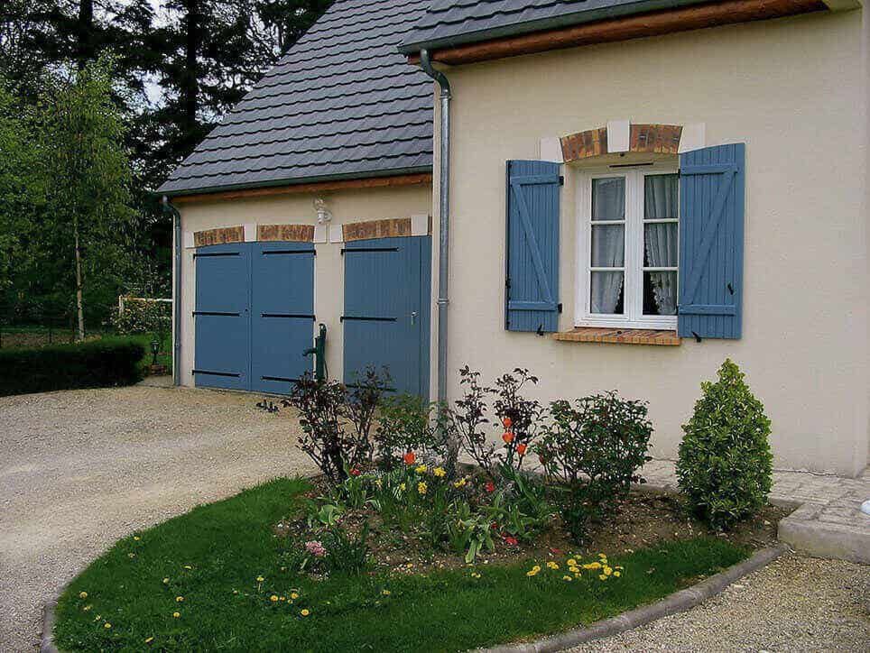 Une maison de couleur claire avec des volets bleus, un parterre de jardin et une allée de gravier.