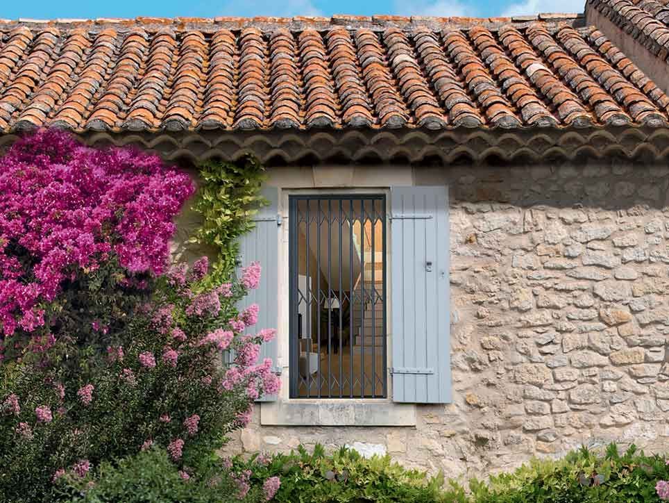 Fenêtre d'un immeuble en pierre aux volets bleus, couverte d'un toit de tuiles rouges et d'un buisson rose en fleurs.