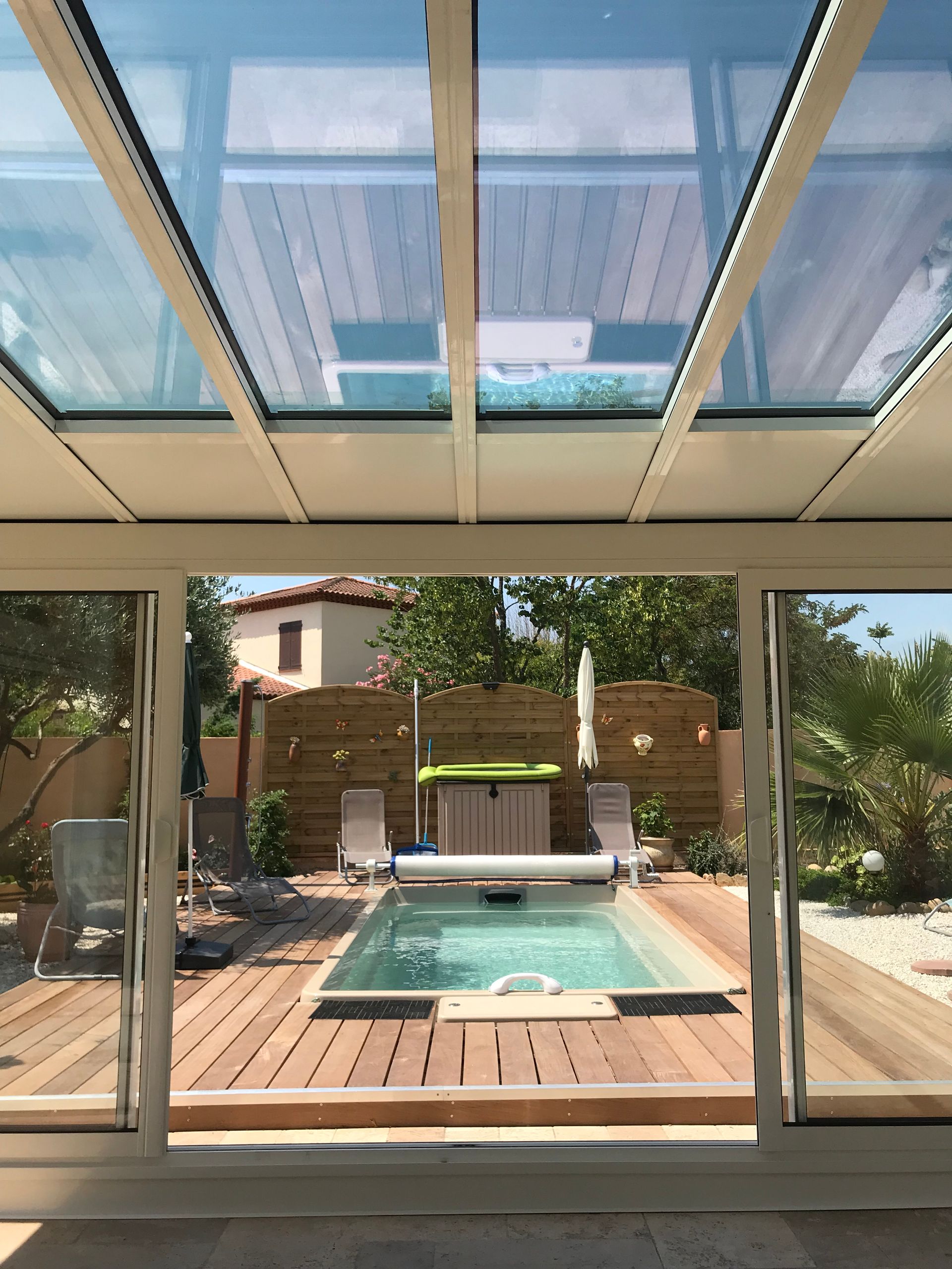 Piscine intérieure avec toit transparent, vue depuis la structure fermée. Terrasse en bois, chaises longues et jardin visibles.