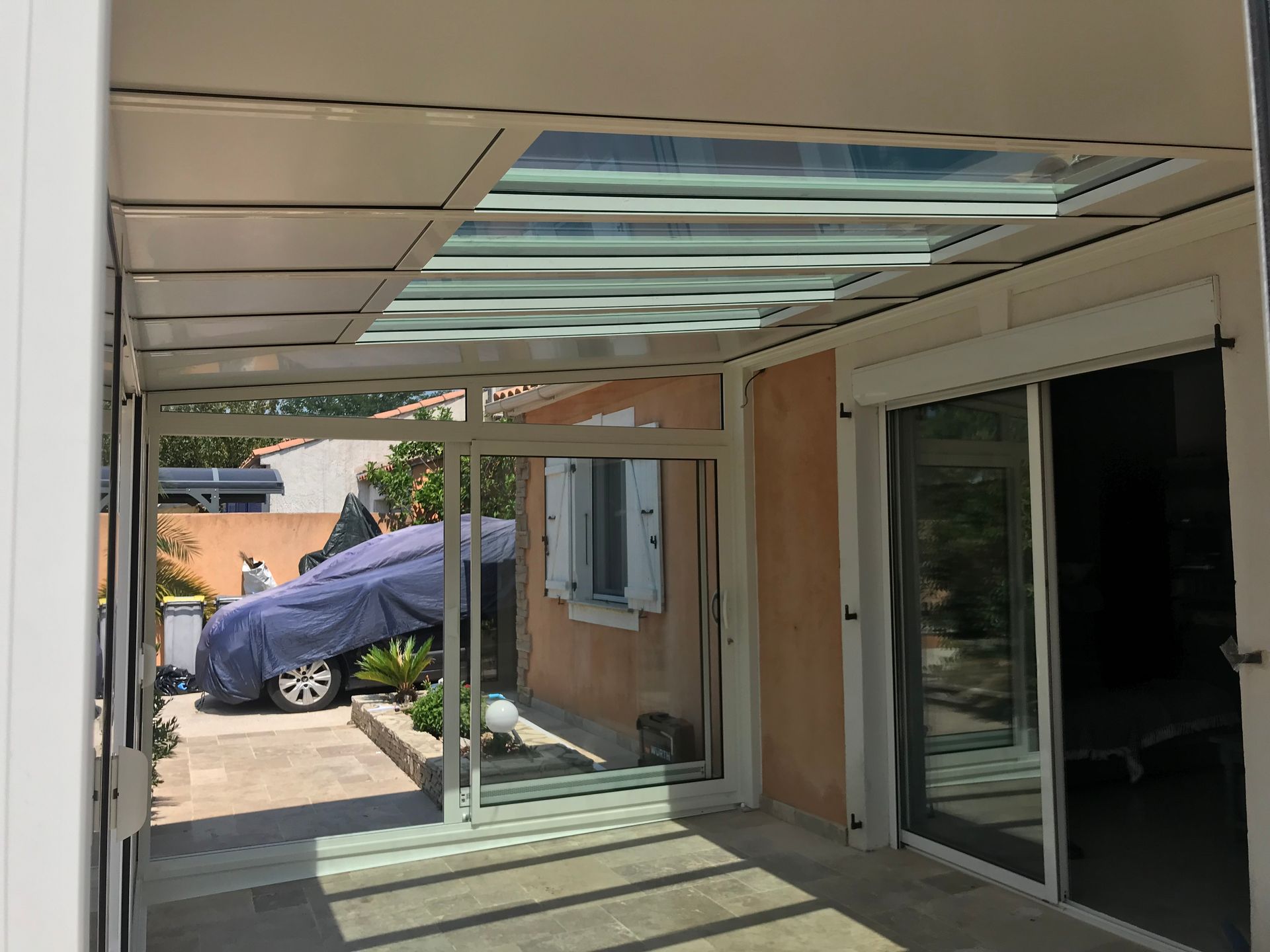 Terrasse extérieure couverte avec une partie de toit en verre, donnant sur une allée et une maison.