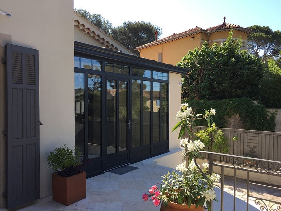 Extérieur d'une maison avec une véranda aux parois de verre, des volets gris et des plantes en pot sur un patio.