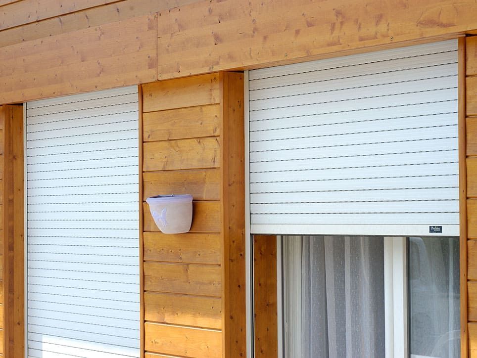 Volets roulants blancs sur un bâtiment en bois, une petite jardinière est montée sur le mur.