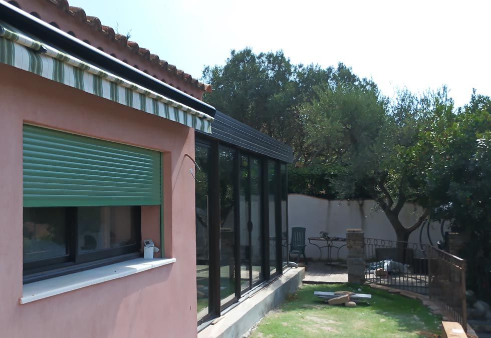 Vue extérieure d'une maison rose avec un volet vert et une véranda aux parois de verre, située dans un espace extérieur ensoleillé.