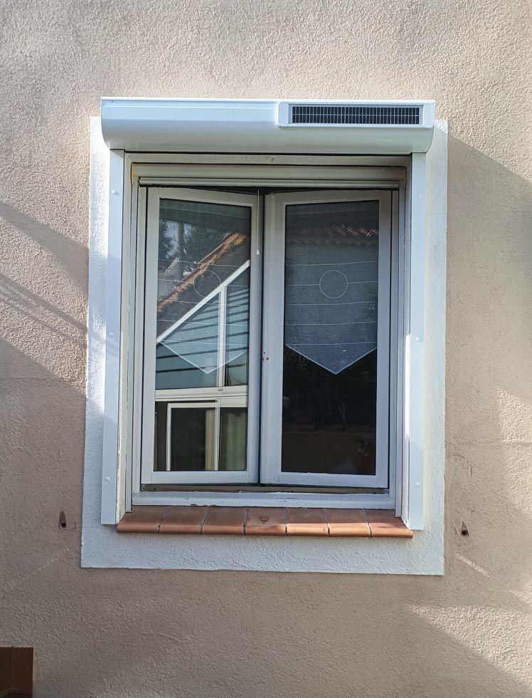 Fenêtre avec un cadre blanc, partiellement ouverte pour révéler l'intérieur, et un volet roulant blanc fermé au-dessus.