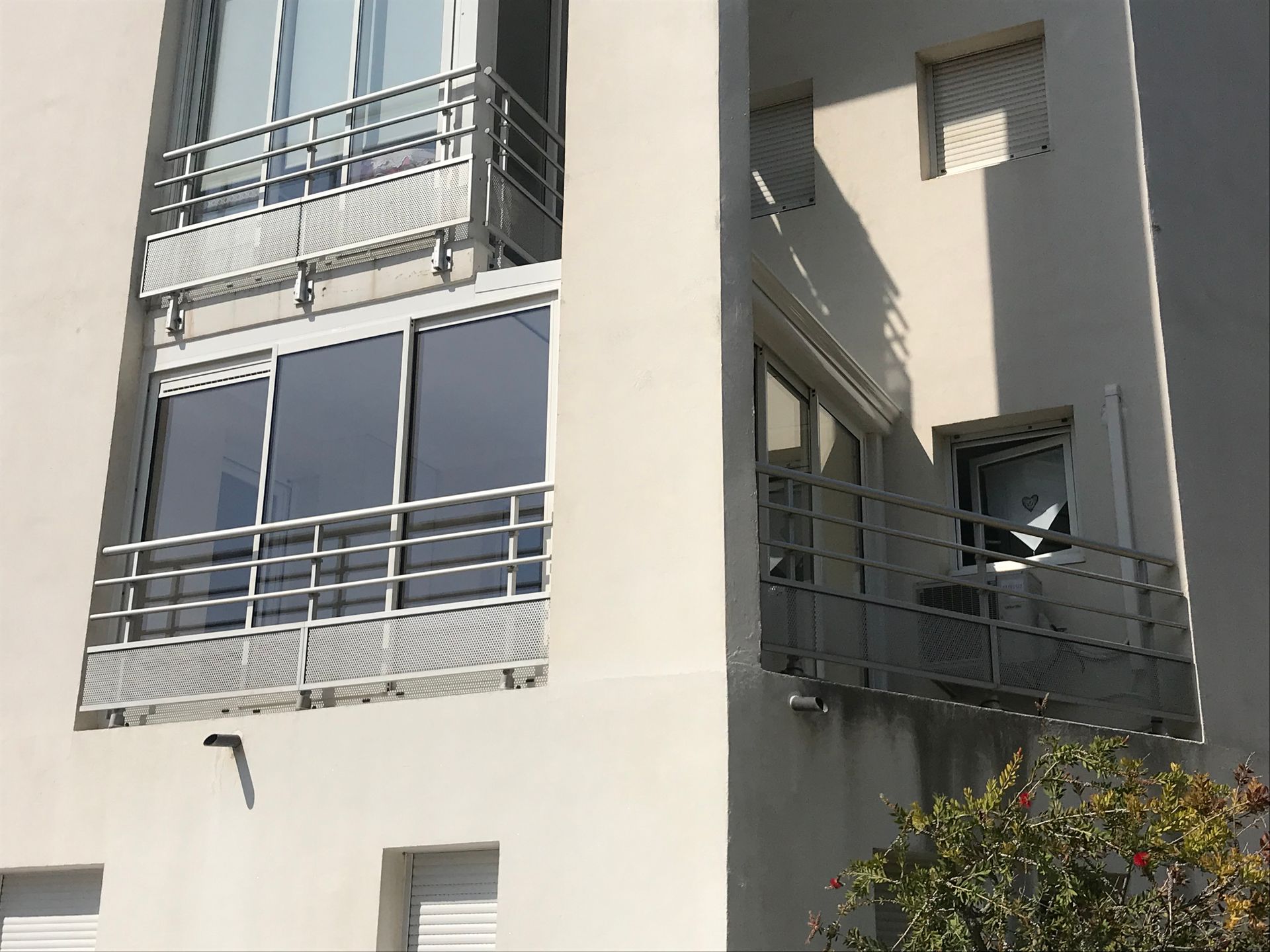 Extérieur d'un bâtiment blanc avec balcons et balustrades métalliques, arbres en contrebas.