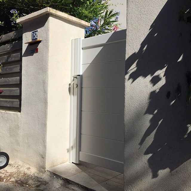 Portail blanc dans un mur en stuc beige avec une boîte aux lettres ; la lumière du soleil projette des ombres.