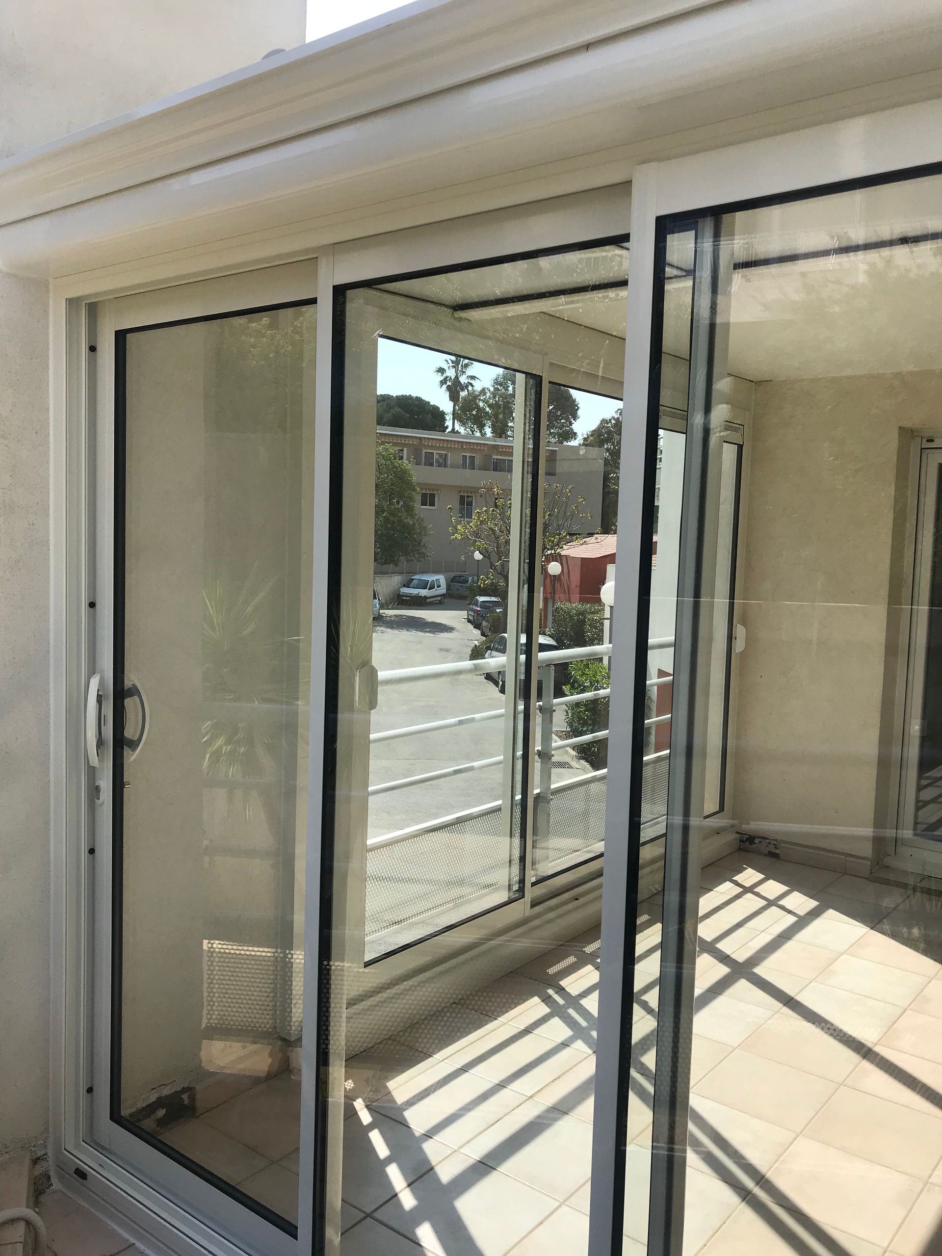 Des portes coulissantes en verre s'ouvrent sur un balcon avec vue sur une rue, des arbres et des bâtiments ; garniture blanche.