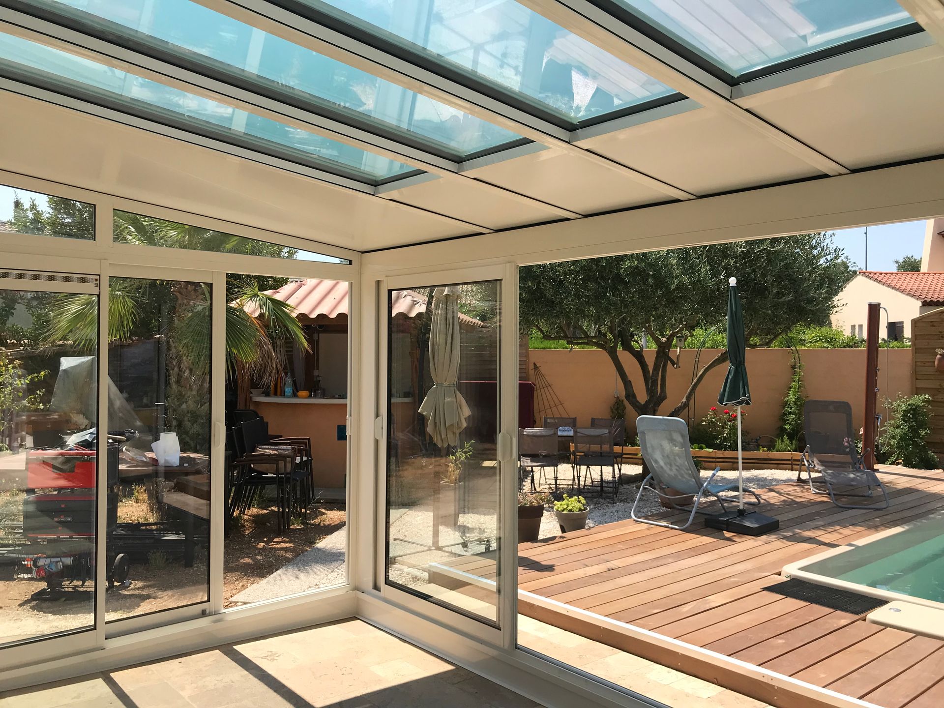 Véranda avec plafond et murs en verre, donnant sur une cour arrière avec terrasse, piscine et arbres.