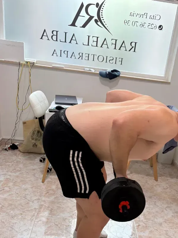Un hombre levanta una mancuerna en un gimnasio, inclinado cerca de una ventana con la inscripción 