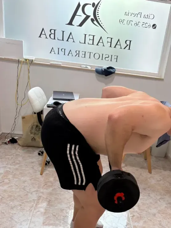Un hombre levanta una mancuerna en un gimnasio, inclinado cerca de una ventana con la inscripción 