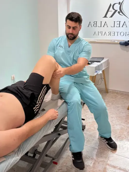 Un fisioterapeuta con uniforme azul examina la pierna flexionada de un paciente en una camilla de tratamiento.