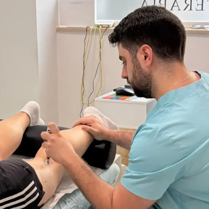 Un fisioterapeuta atiende la rodilla de un paciente en una clínica, con la pierna apoyada sobre una mesa acolchada.