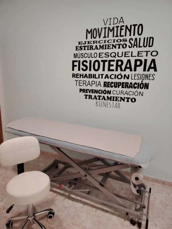 Camilla y taburete para tratamiento de fisioterapia en una clínica, con palabras en español en la pared.
