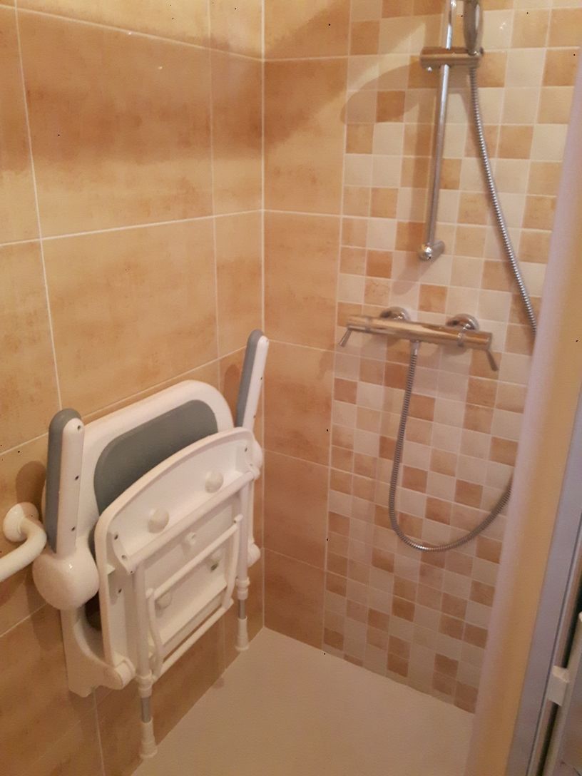Siège de douche rabattable fixé au mur carrelé de la salle de bain. Pommeau de douche et robinet visibles.