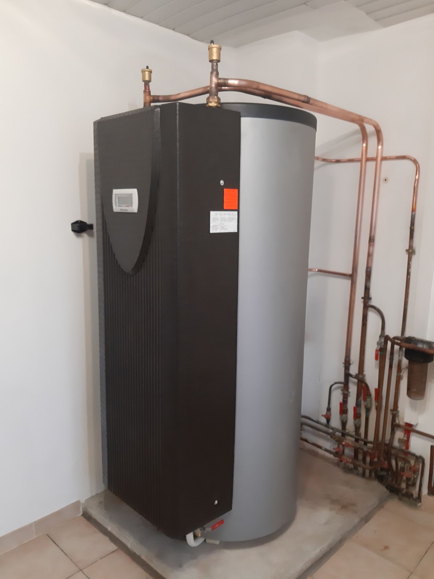Un grand chauffe-eau avec des tuyaux en cuivre contre un mur blanc.