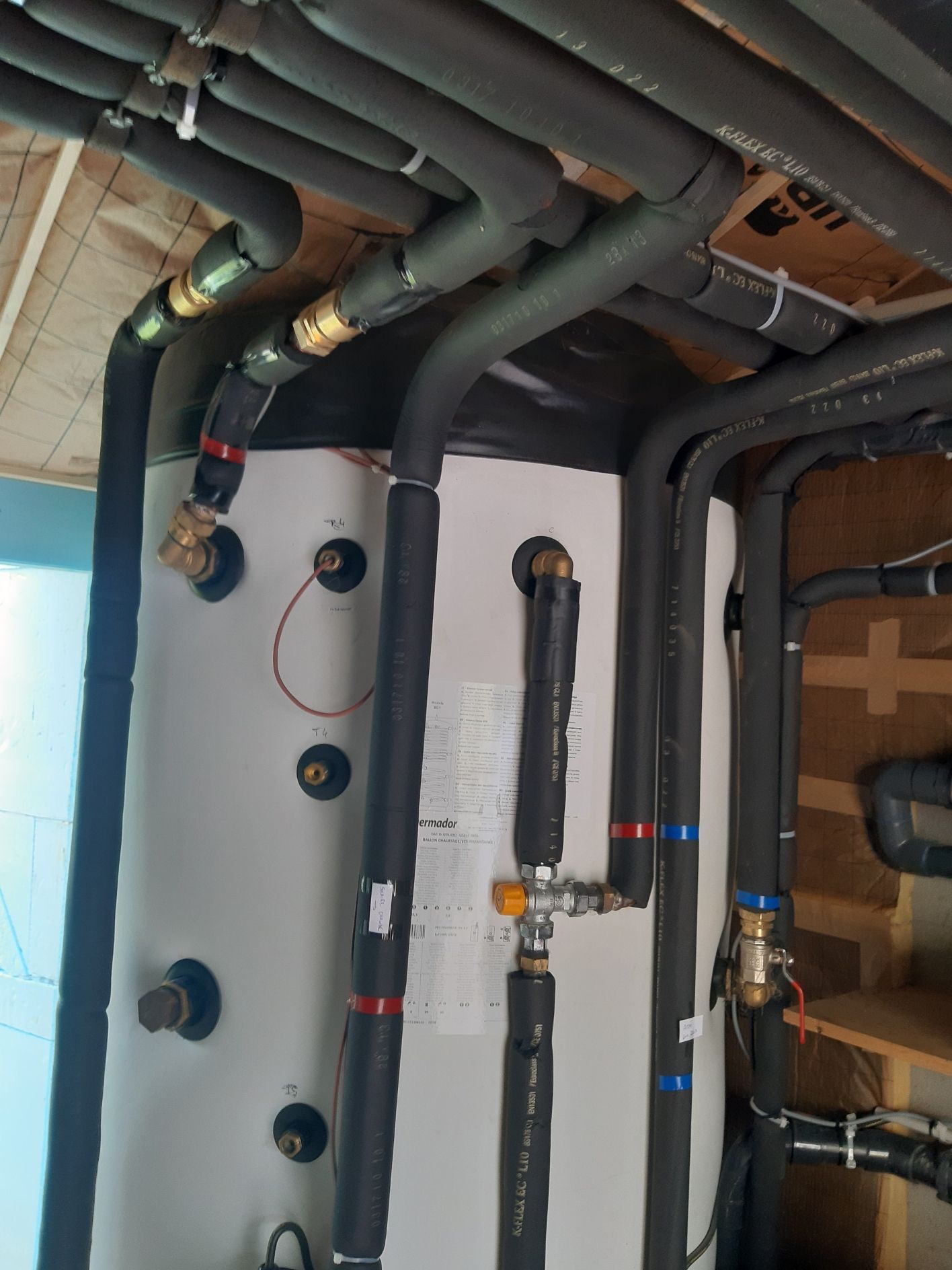 Tuyaux et raccords isolés avec de la mousse noire, reliés à un réservoir de chauffe-eau blanc, à l'intérieur d'un local.