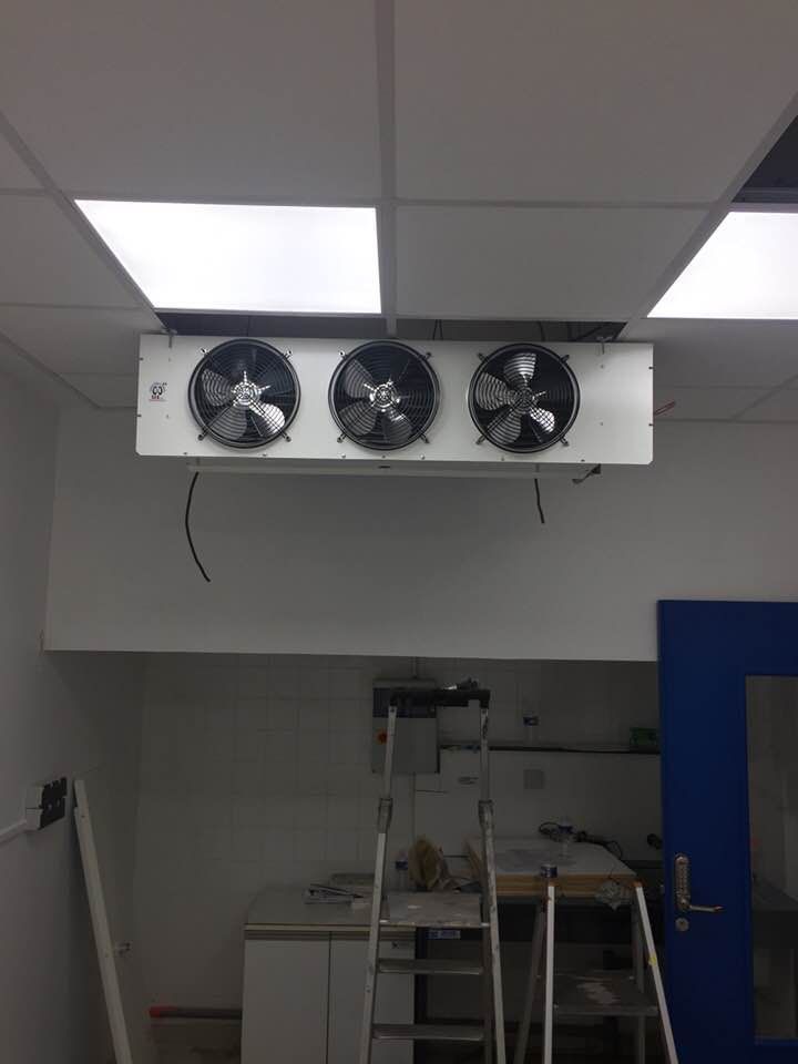 Unité de refroidissement industrielle blanche fixée au plafond, équipée de trois ventilateurs, installée au-dessus d'un espace de travail.