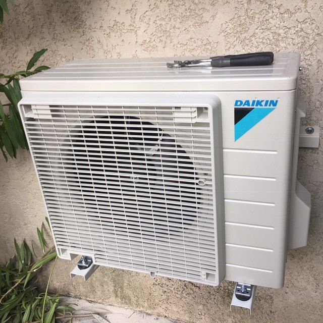 Un climatiseur split extérieur Daikin blanc, fixé sur un mur beige, avec une paire de pinces posée dessus.