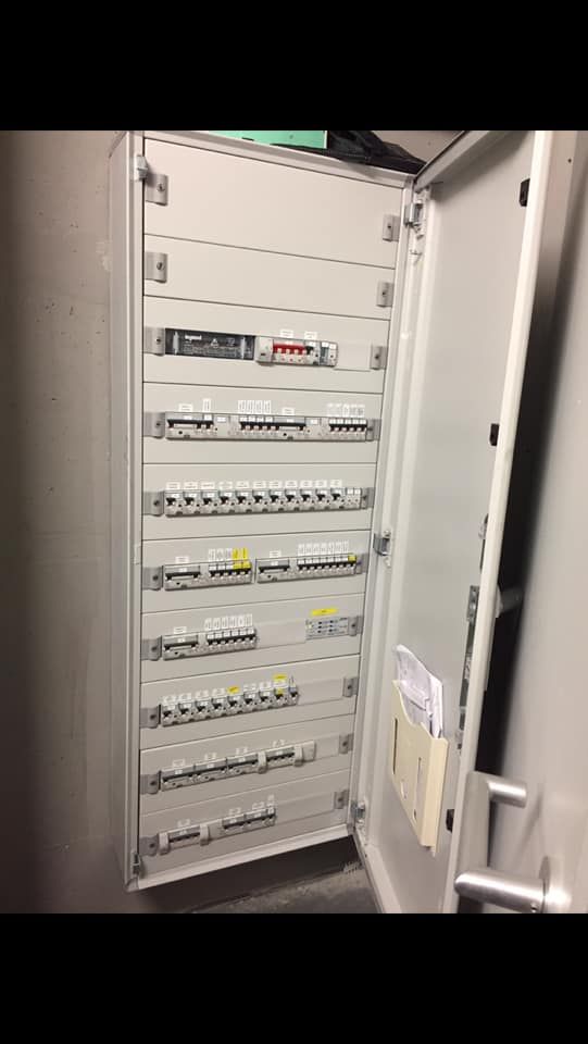 Un tableau de distribution électrique ouvert, gris clair, comportant plusieurs rangées de disjoncteurs et d'interrupteurs.