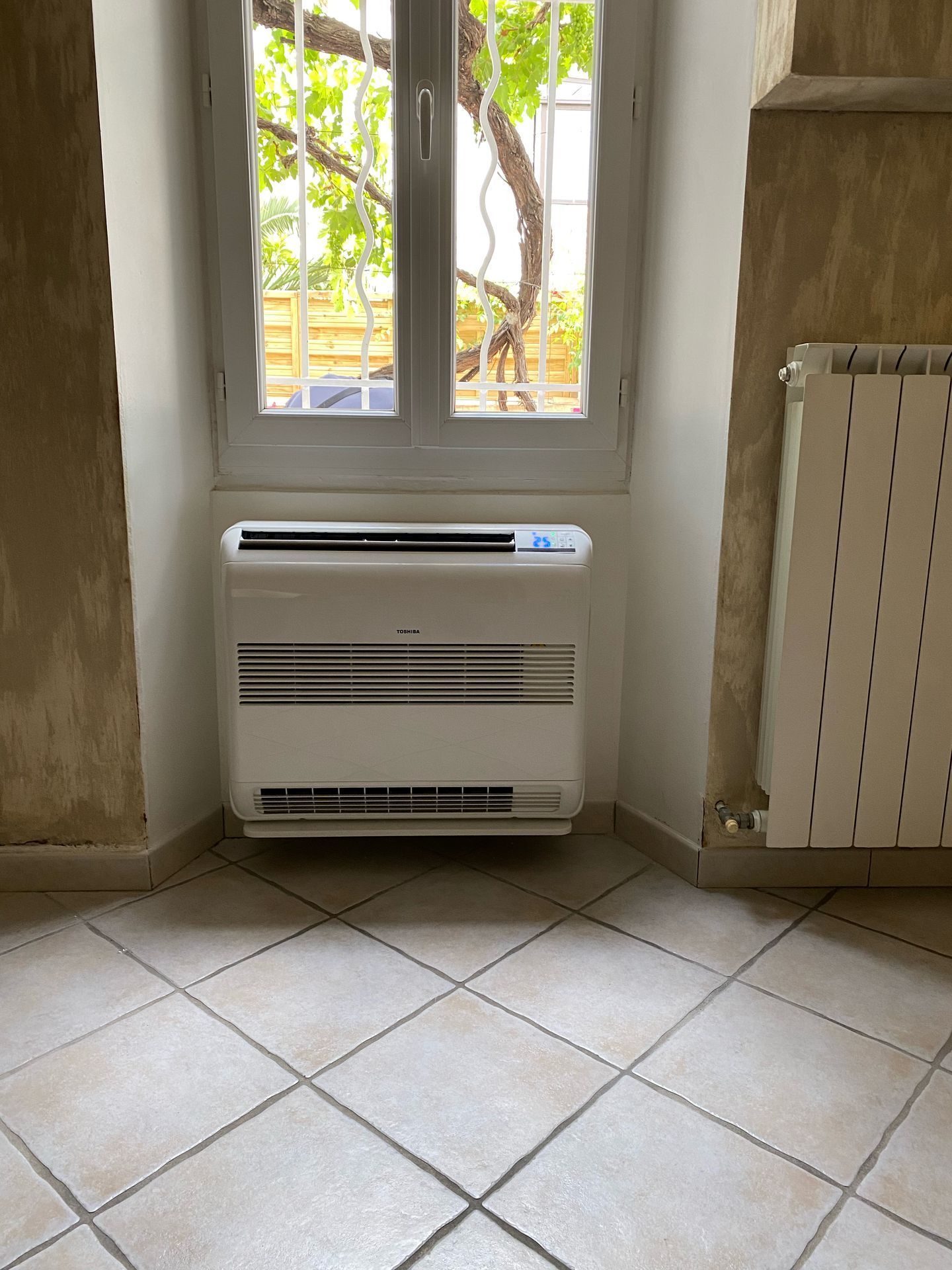 Un climatiseur blanc sur pied, installé sous une fenêtre dans une pièce au sol carrelé et équipée d'un radiateur mural.