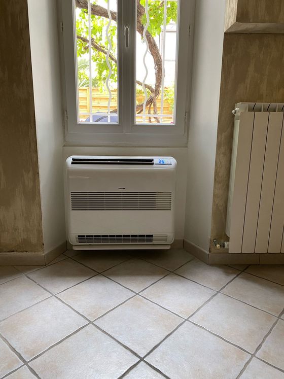 Un climatiseur blanc sur pied, installé sous une fenêtre dans une pièce au sol carrelé et équipée d'un radiateur mural.