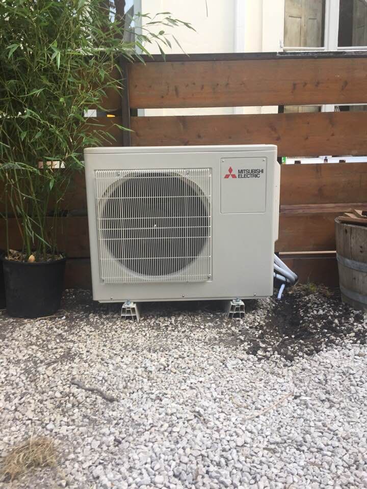 Un climatiseur extérieur Mitsubishi est installé sur un sol gravillonné, devant une clôture en bois et un bambou en pot.