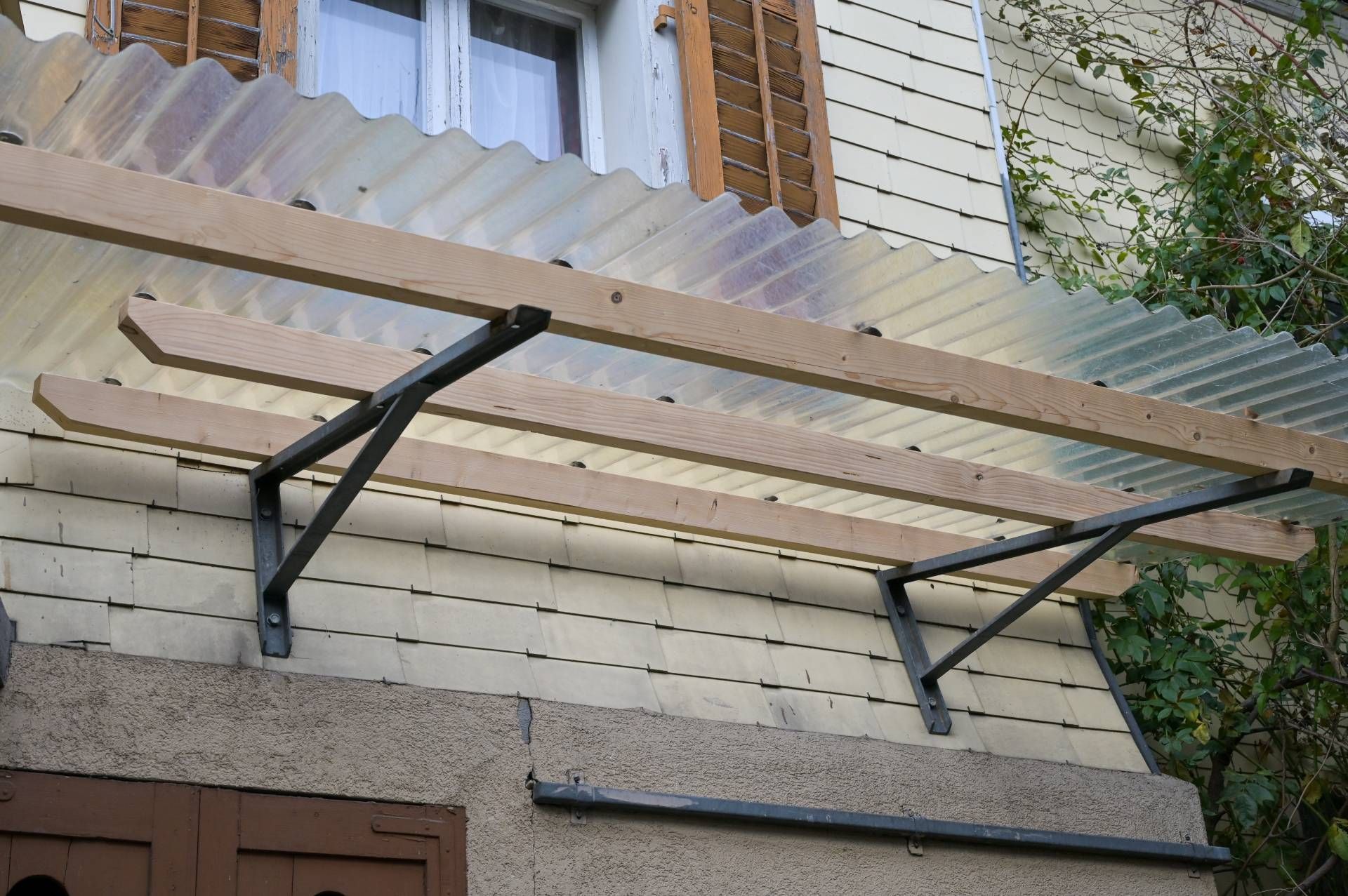 Über einer Aussentür befindet sich ein transparentes Wellblechdach, das von Holzbalken und Metallhalterungen getragen wird.