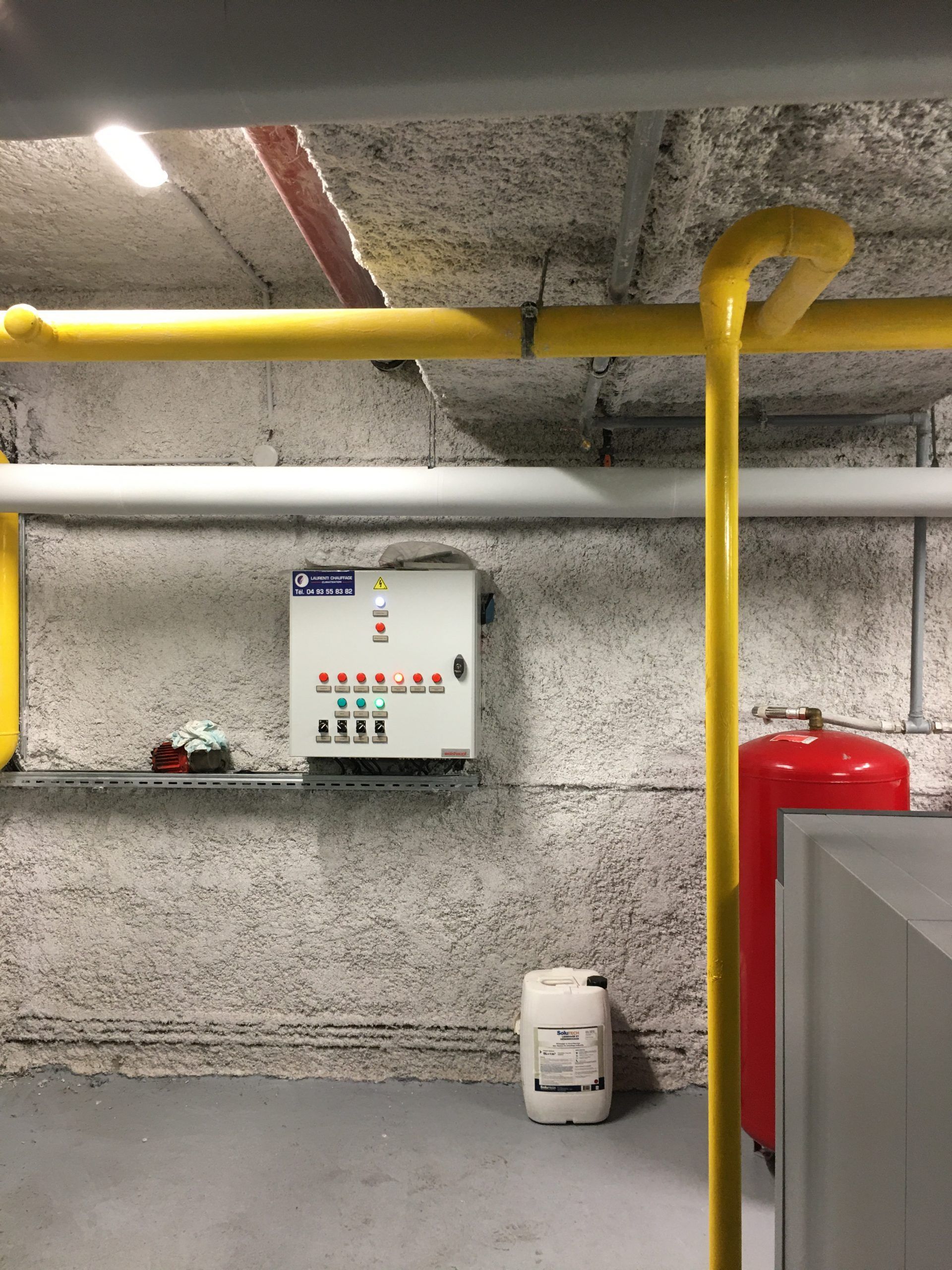Tableau électrique et plomberie dans une buanderie. Tuyaux jaunes et blancs sur un plafond et un mur texturés.