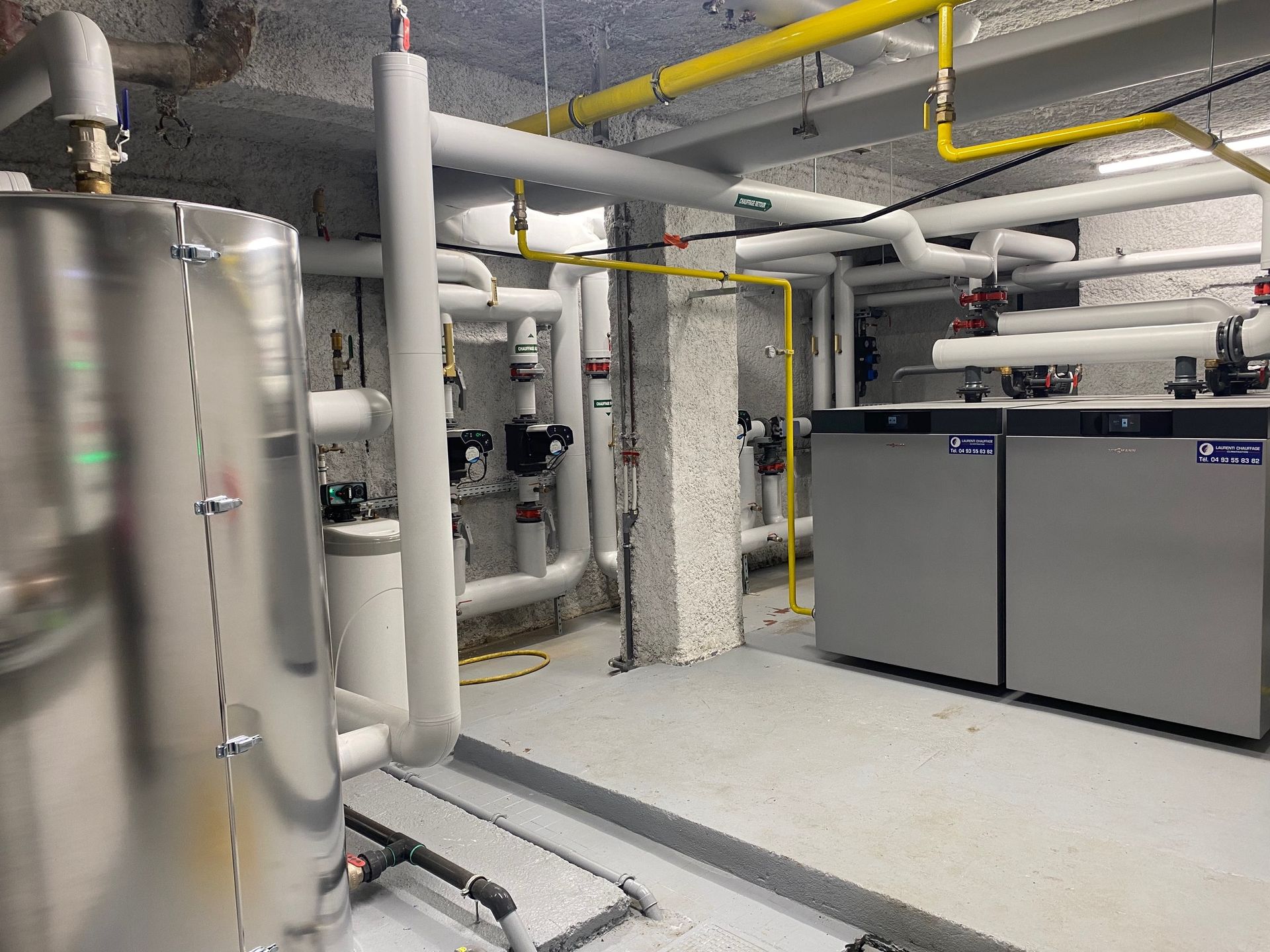 Local technique avec tuyauterie, réservoirs et deux chaudières grises. Tuyaux jaunes et blancs contre les murs en béton.
