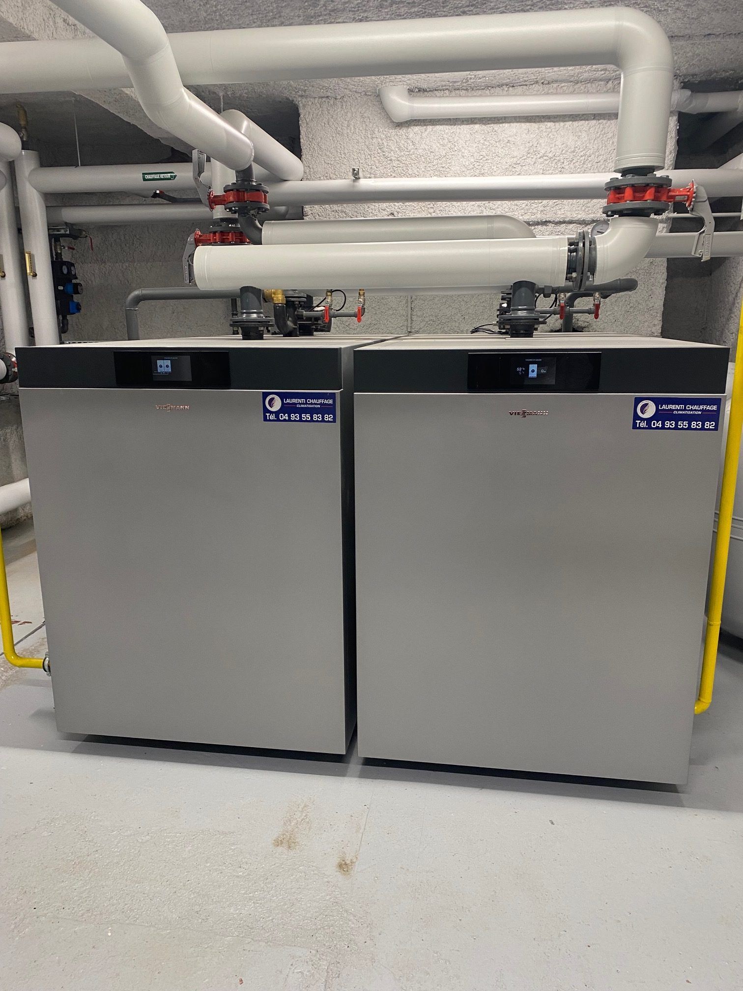 Deux chaudières argentées dans une buanderie avec des tuyaux blancs au plafond.