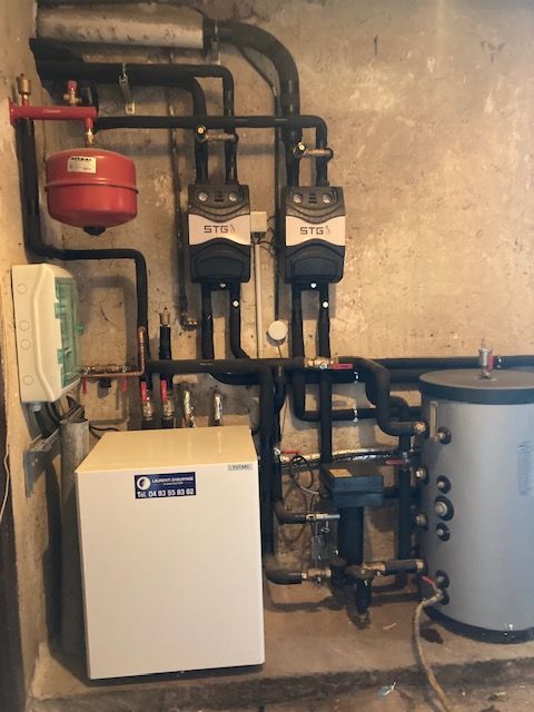 Composants du système de chauffage fixés sur un mur en béton : chaudière, pompes, vase d’expansion, chauffe-eau et tuyauterie.