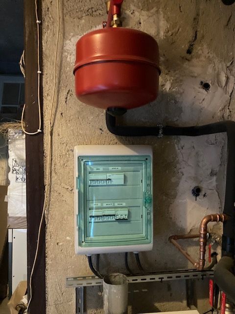 Vase d'expansion rouge au-dessus d'un tableau électrique blanc, sur un mur en béton brut avec des tuyaux.