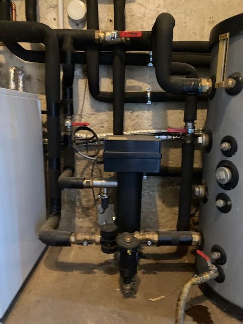 Système de tuyauterie avec tuyaux noirs isolés, vannes et chauffe-eau cylindrique contre un mur texturé.