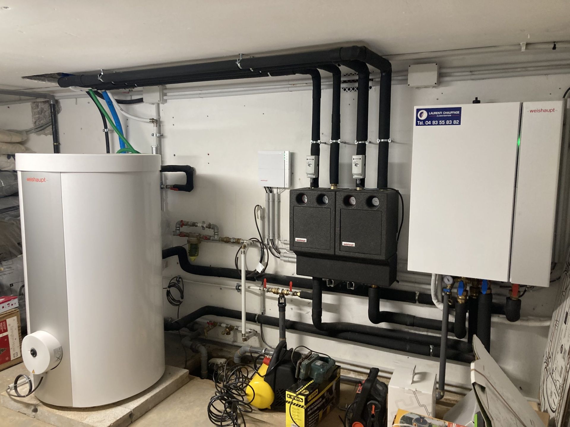 Un chauffe-eau cylindrique blanc et un système de chauffage, ventilation et climatisation se trouvent dans un sous-sol. Des tuyaux noirs et des câbles électriques sont visibles.