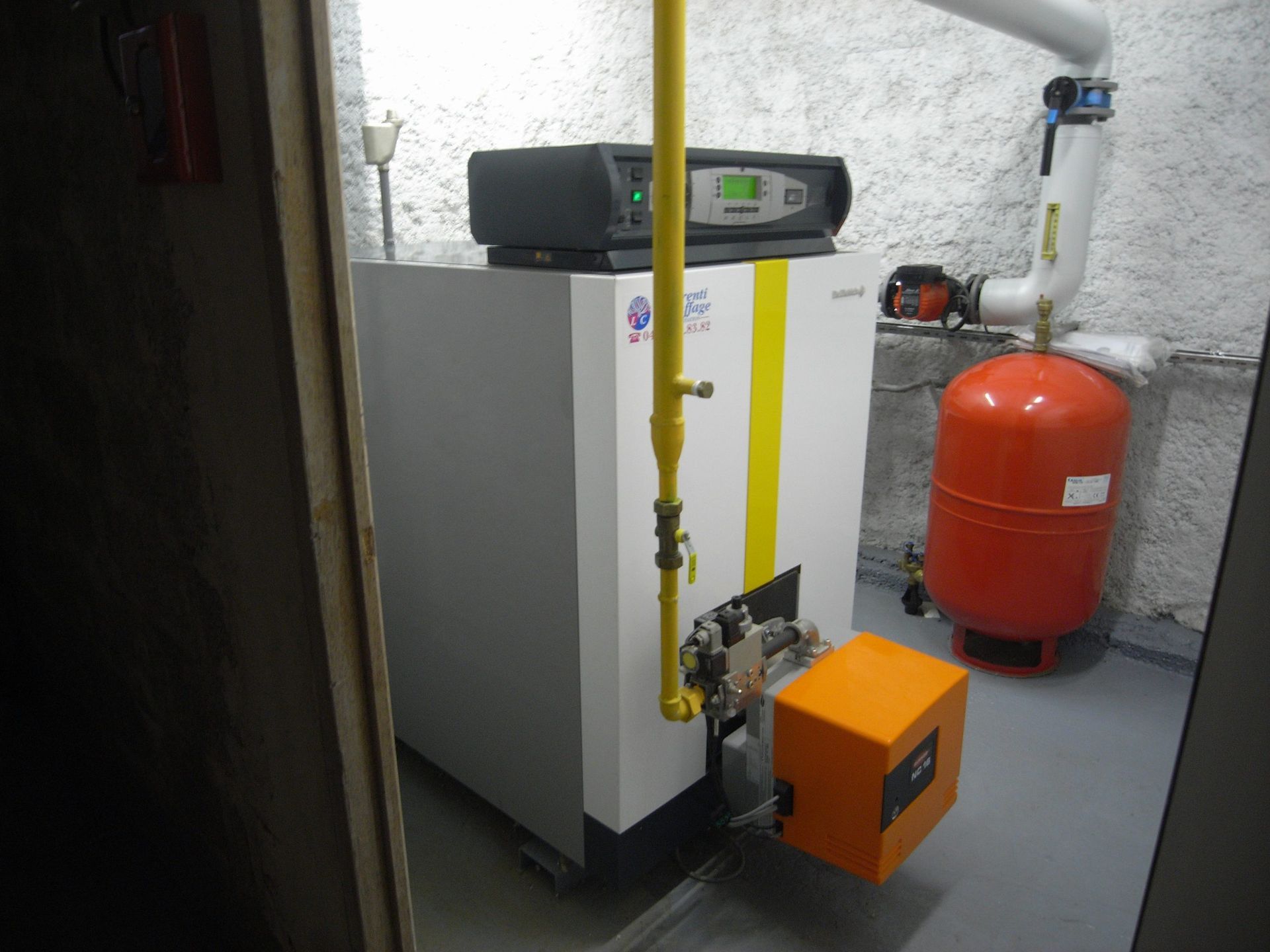 Une chaudière blanche et jaune dans une buanderie. Un vase d'expansion orange se trouve à proximité.