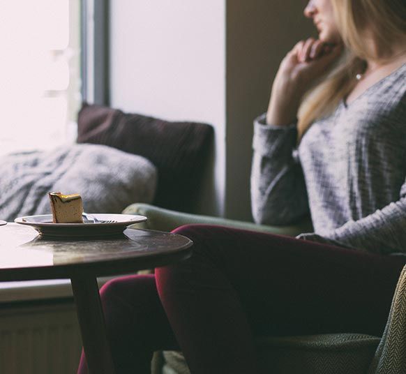 Une femme assise près d'une fenêtre, la main près du visage, avec une part de gâteau sur une table.