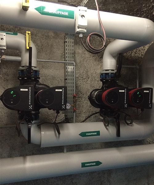 Deux pompes Grundfos sombres, reliées à des tuyaux gris avec des étiquettes vertes et un câblage électrique, sont fixées au mur.