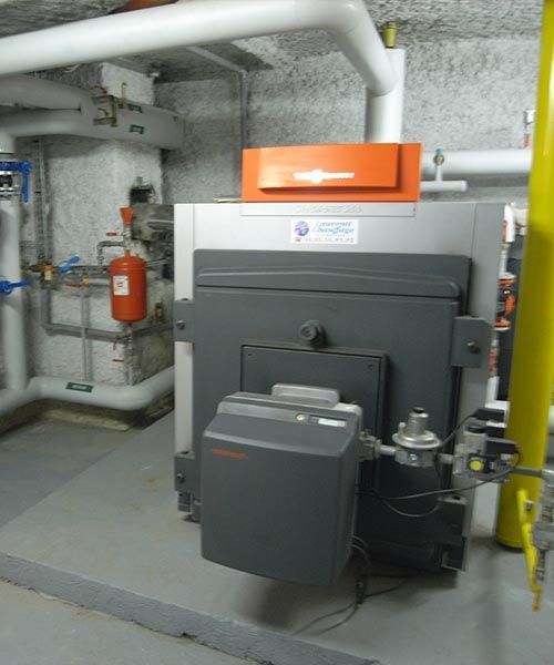 Grosse chaudière industrielle en sous-sol. Extérieur en métal gris, dessus orange, tuyauterie raccordée.