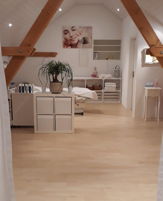 Ein minimalistischer Behandlungsraum im Dachgeschoss mit hellem Holzboden, einer Massageliege und weißen Regalen.