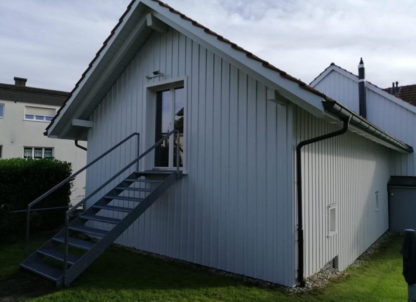 Eine helle Hausfassade mit einer Metalltreppe, die zu einem erhöhten Eingang hinaufführt.