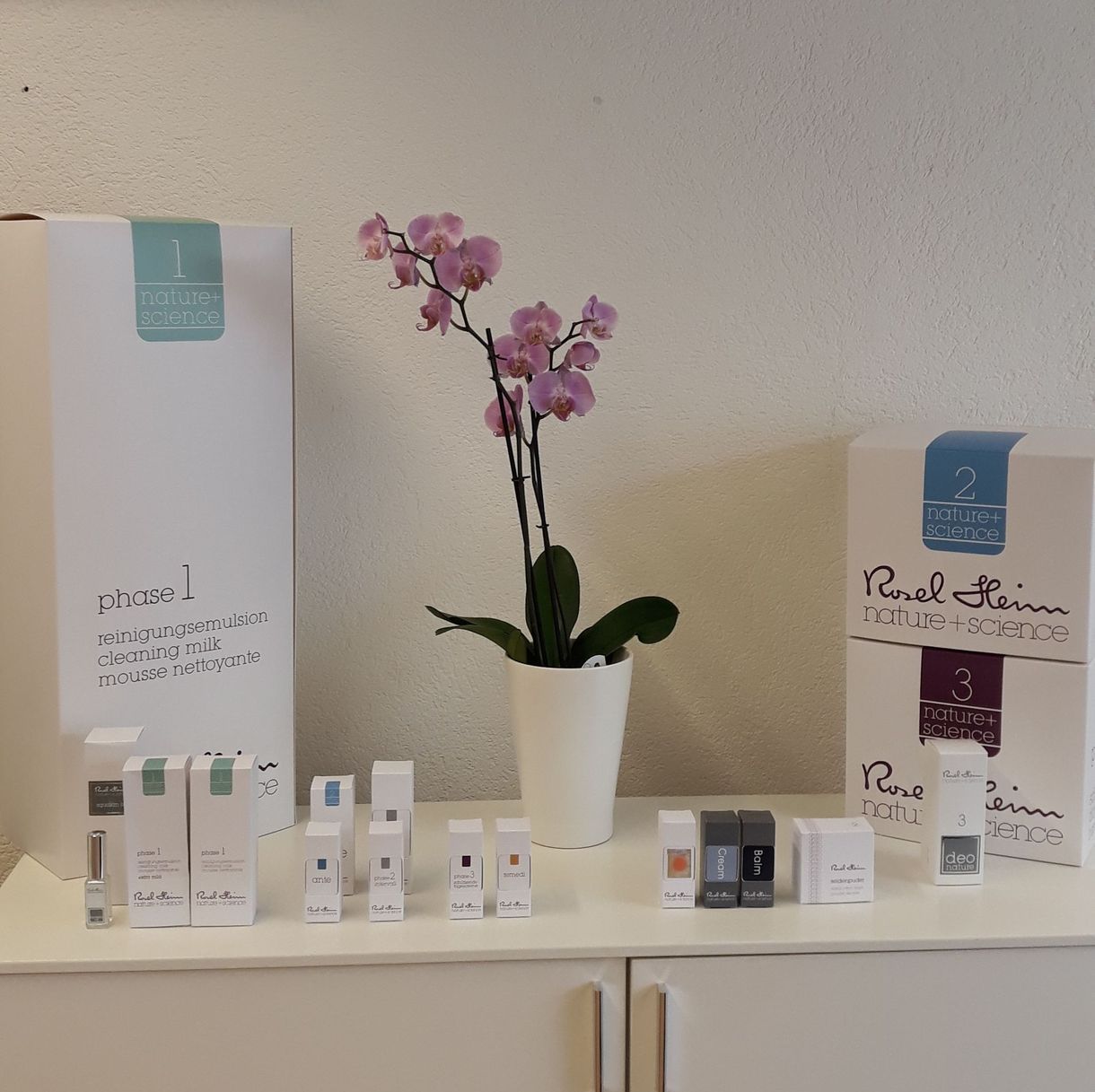 Auf einem weißen Regal sind Hautpflegeprodukte und -verpackungen arrangiert, in deren Mitte eine rosa Orchidee im Topf steht.