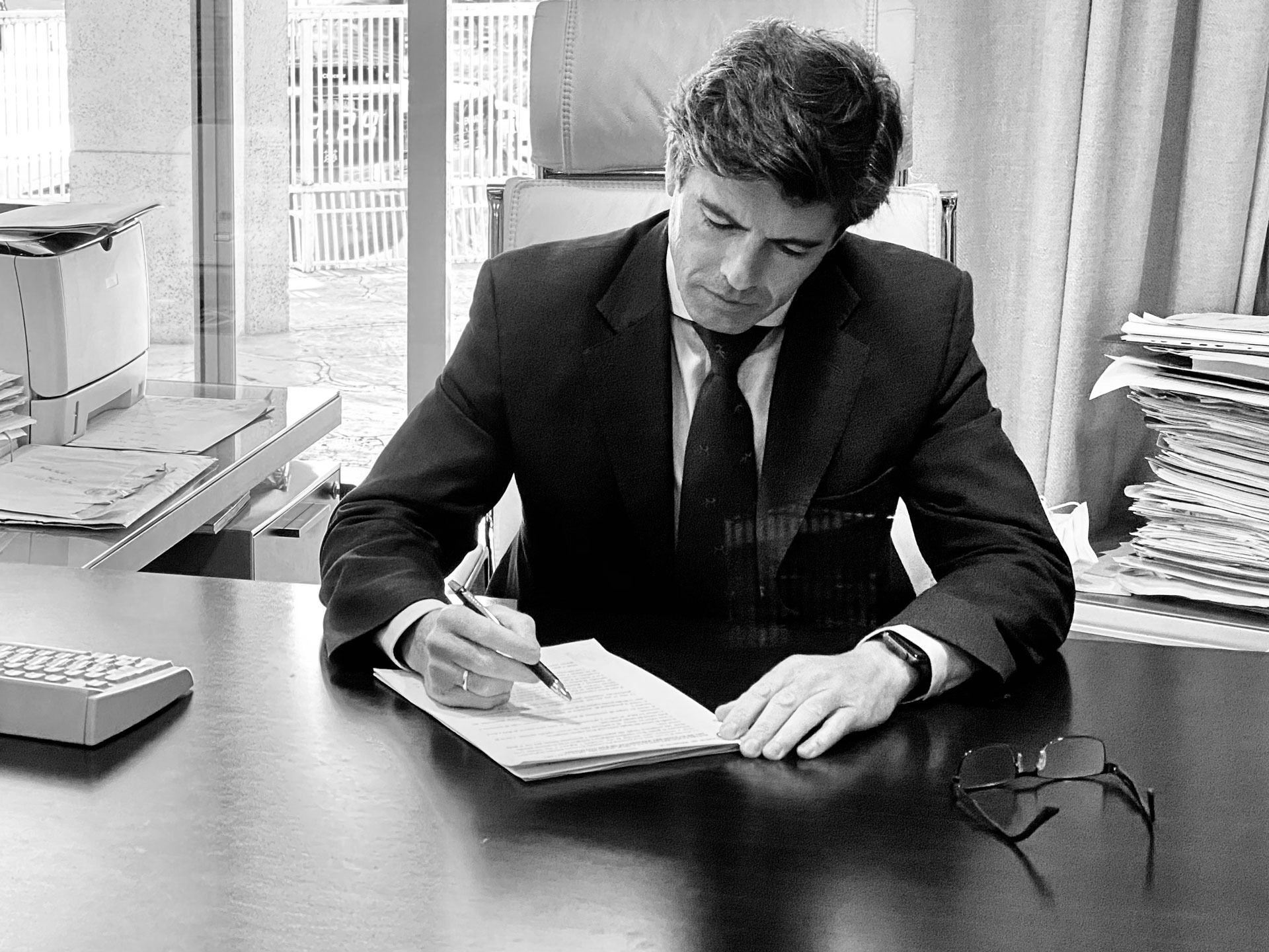 Hombre de traje escribiendo en un escritorio, rodeado de material de oficina y papeles.