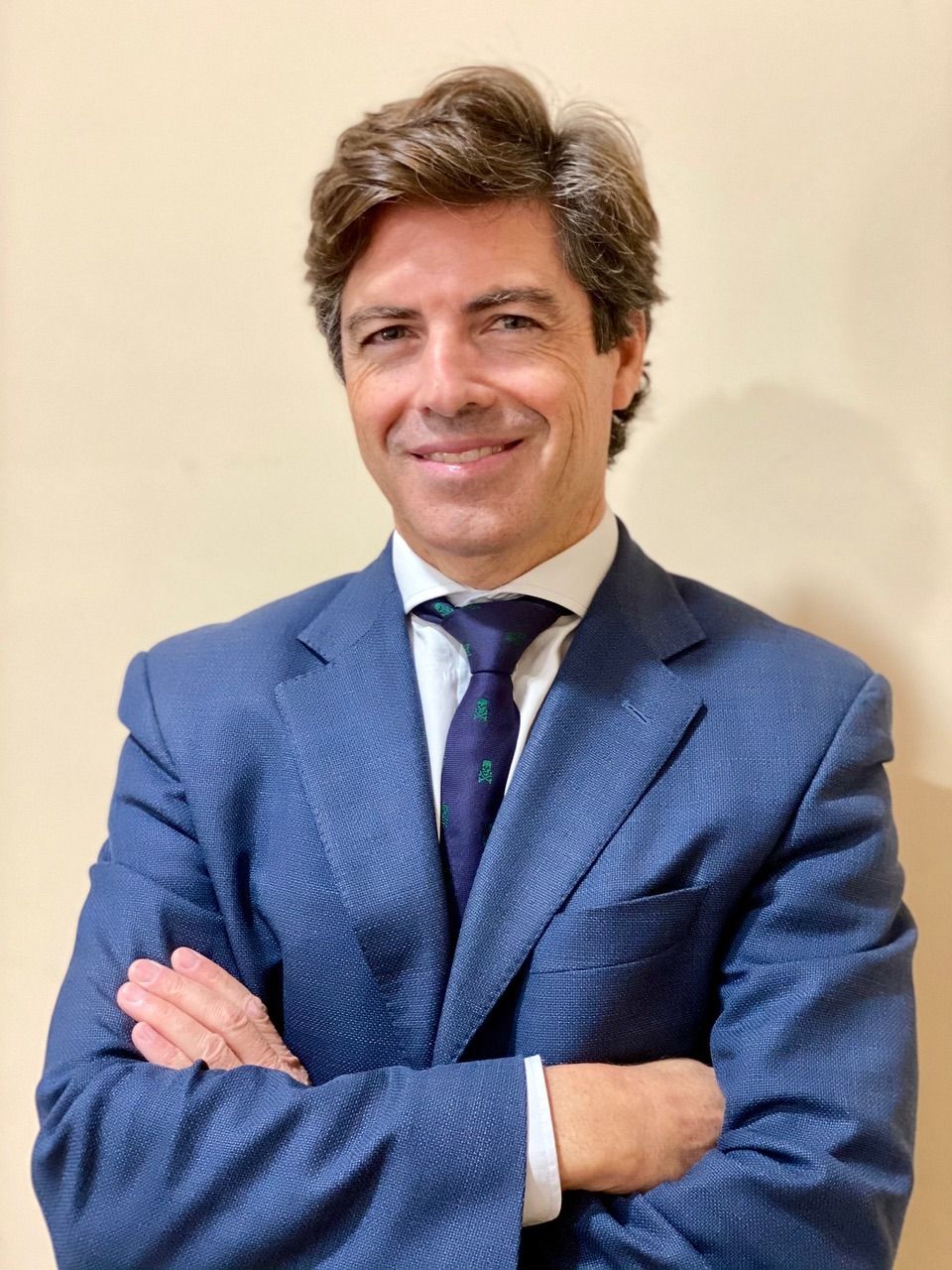 Hombre de traje azul con los brazos cruzados, sonriendo frente a una pared neutra.