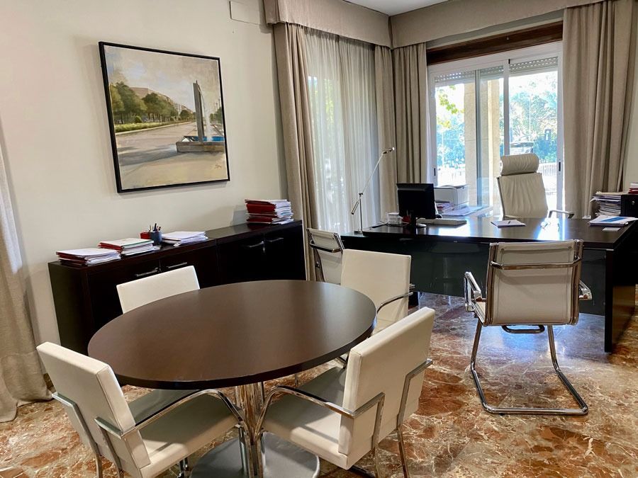 Oficina con escritorio oscuro, mesa redonda y sillas claras; obras de arte enmarcadas y ventana grande.