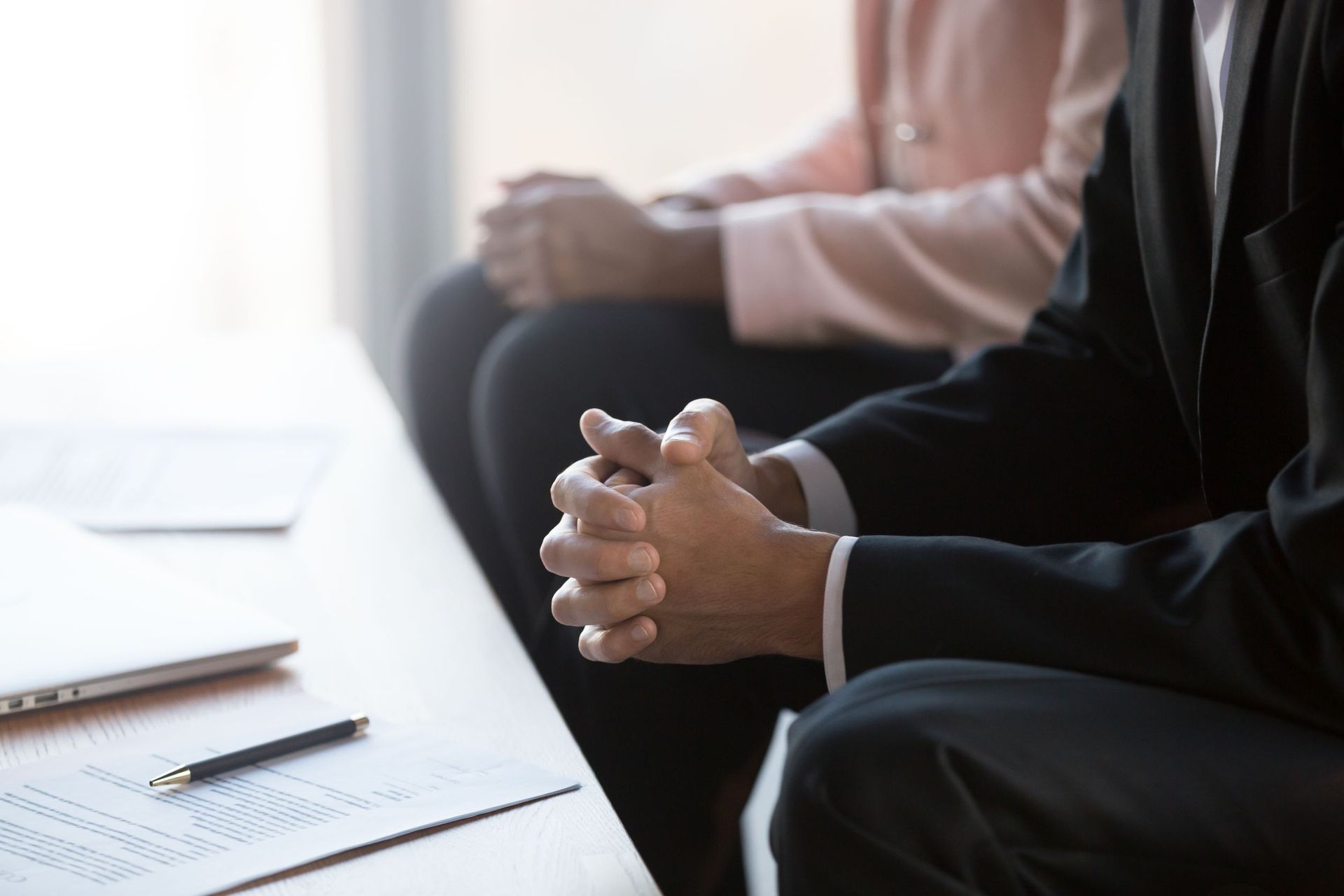 Dos personas sentadas, con las manos entrelazadas, escuchando atentamente en una reunión o entrevista. Escritorio con documentos delante.