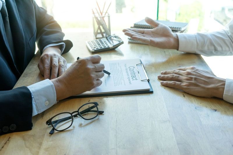 Dos personas en una mesa; una firma un contrato, mientras la otra gesticula. Sobre la mesa hay vasos y una calculadora.